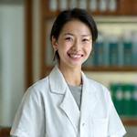 白衣を着用した皮膚科専門医が、優しい笑顔でカメラを見つめている。背後には医学書や医療器具が整然と並んでいる。権威と安心感を醸し出すポートレート。